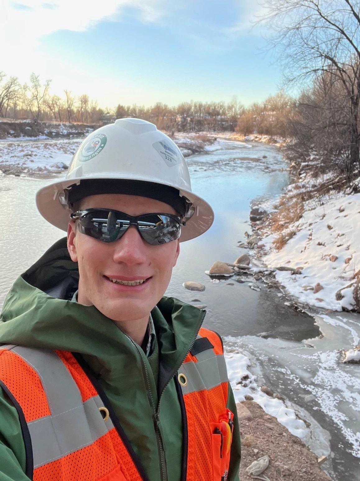 Slade stands near a creek in winter, wearing a high visibility jacket and hard helmet.