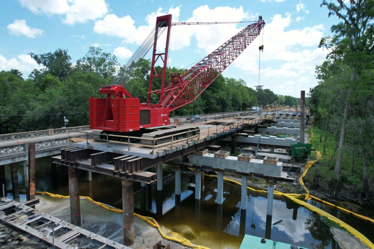 A large crane sitting atop the old bridge lays the new bridge deck over the installed piers.